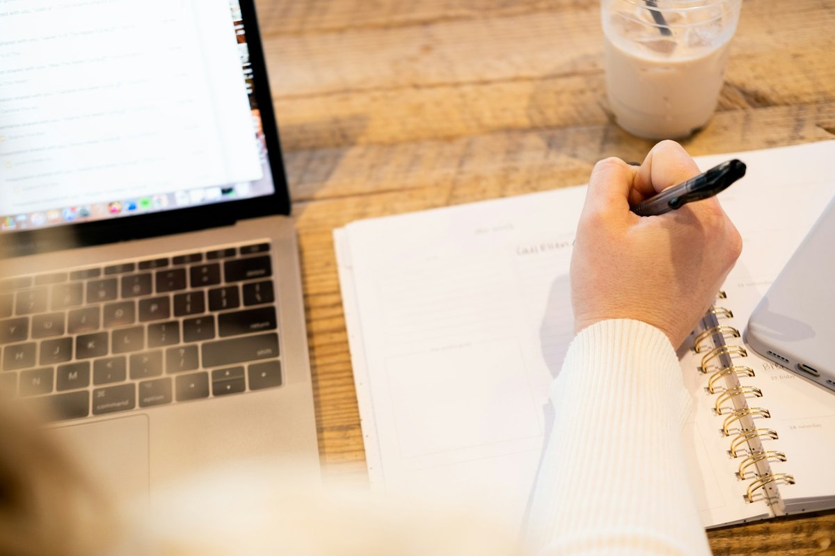 Hand writing in a spiral planner next to an open laptop, preparing a one-page plan before a sales demo call